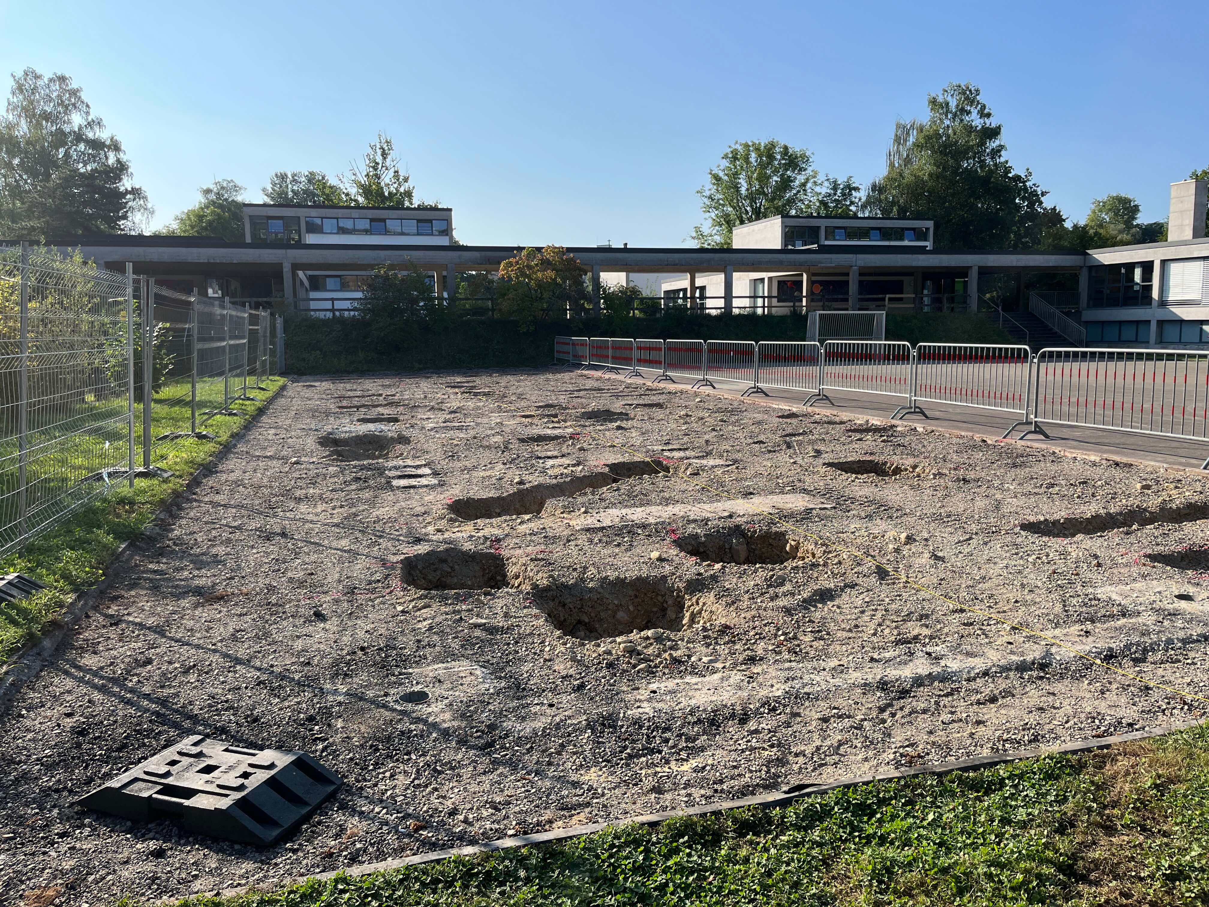 Geplantes Baugelände mit ausgehobenen Löchern und Bauzäunen, umgeben von Grünflächen und Gebäuden im Hintergrund