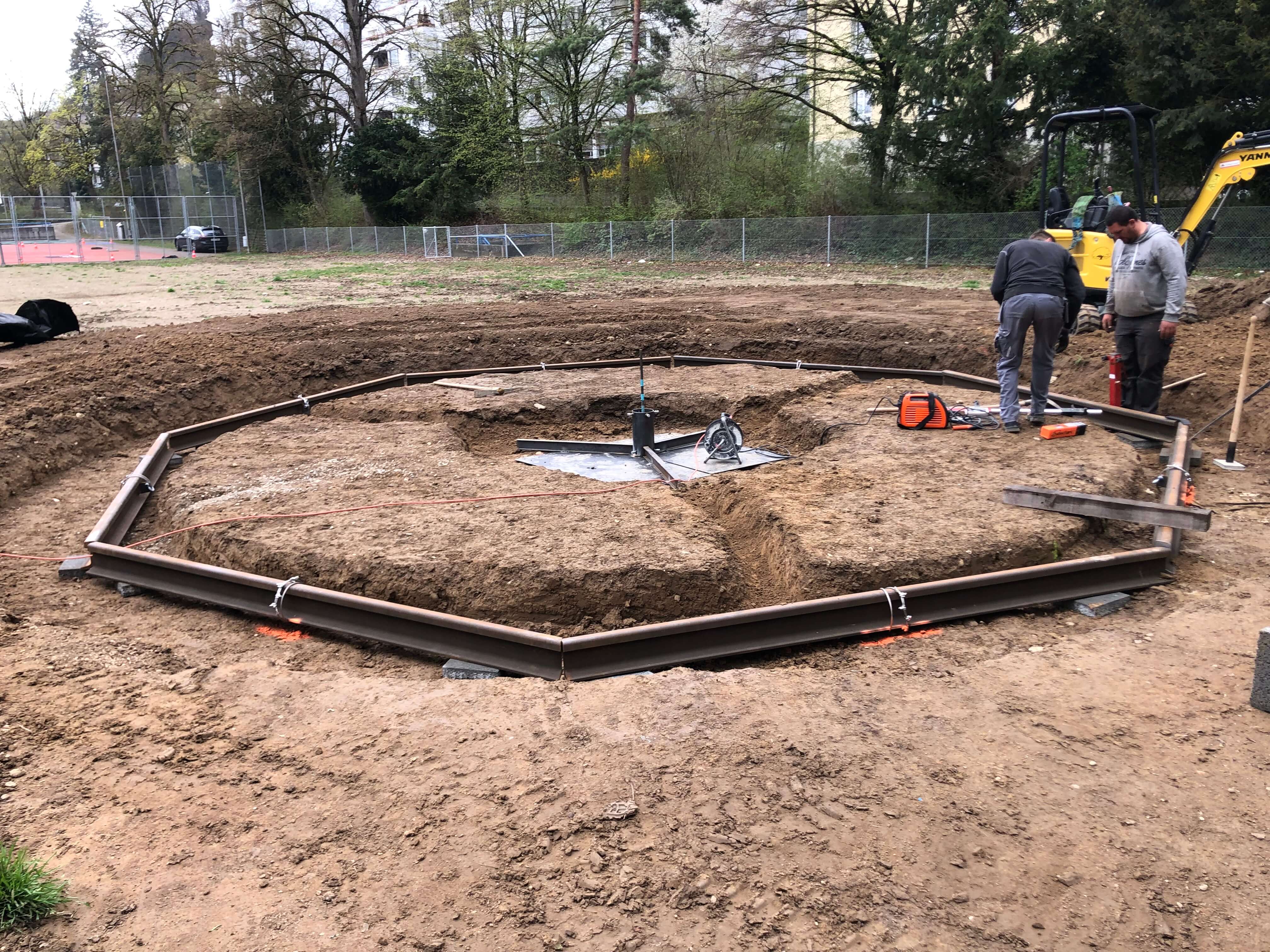 Zwei Bauarbeiter arbeiten an der Konstruktion einer kreisförmigen Kiesbett-Basis auf einer Baustelle im Freien.