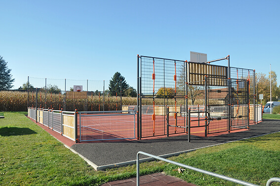 Ein umzäunter Multifunktions-Sportplatz mit Basketballkörben und Fußballtoren, umgeben von Grünflächen.