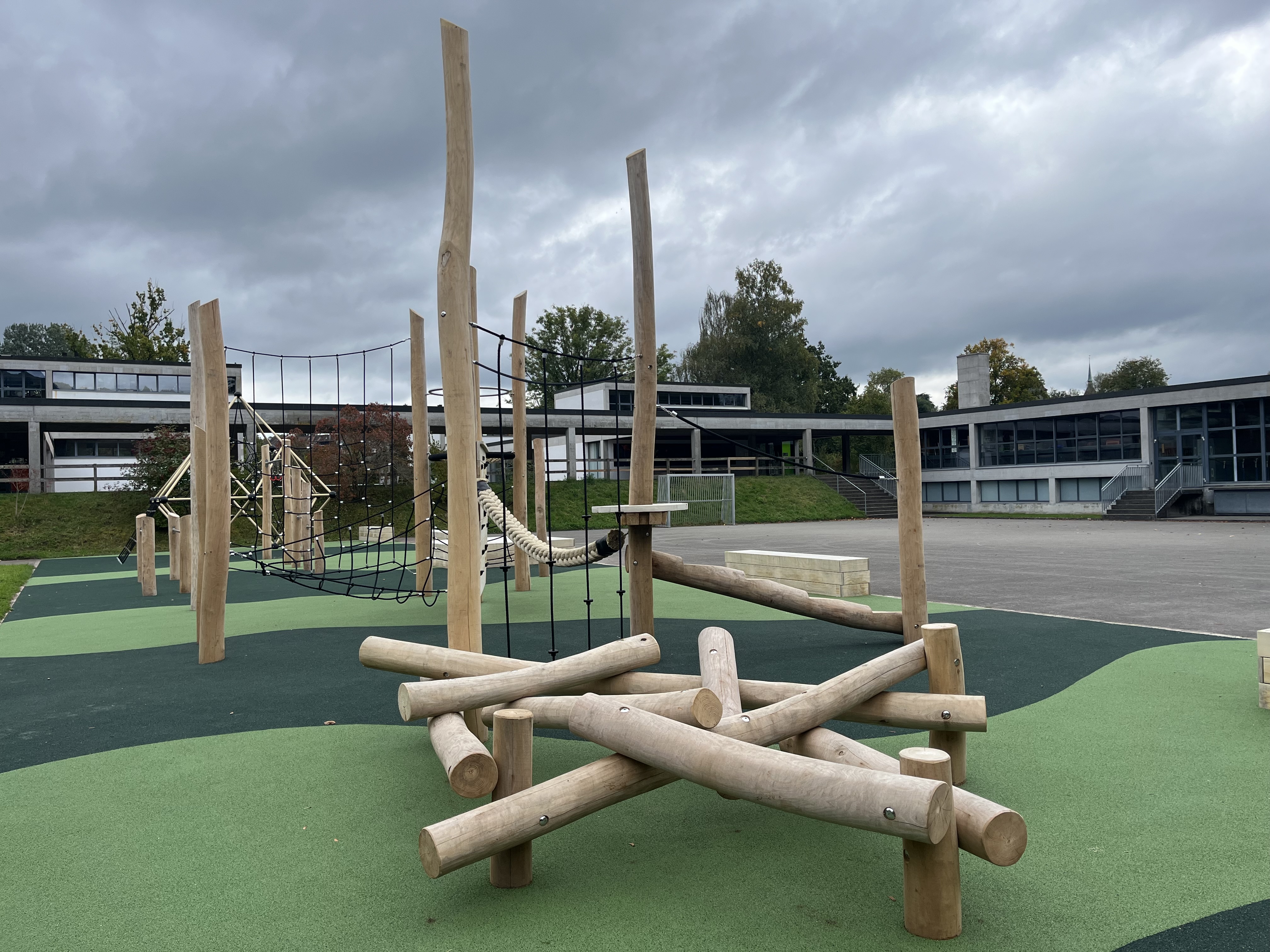 Ein moderner Spielplatz mit einem Klettergerüst aus Holz und Seilen auf einem grünen Gummiboden vor einem Schulgebäude
