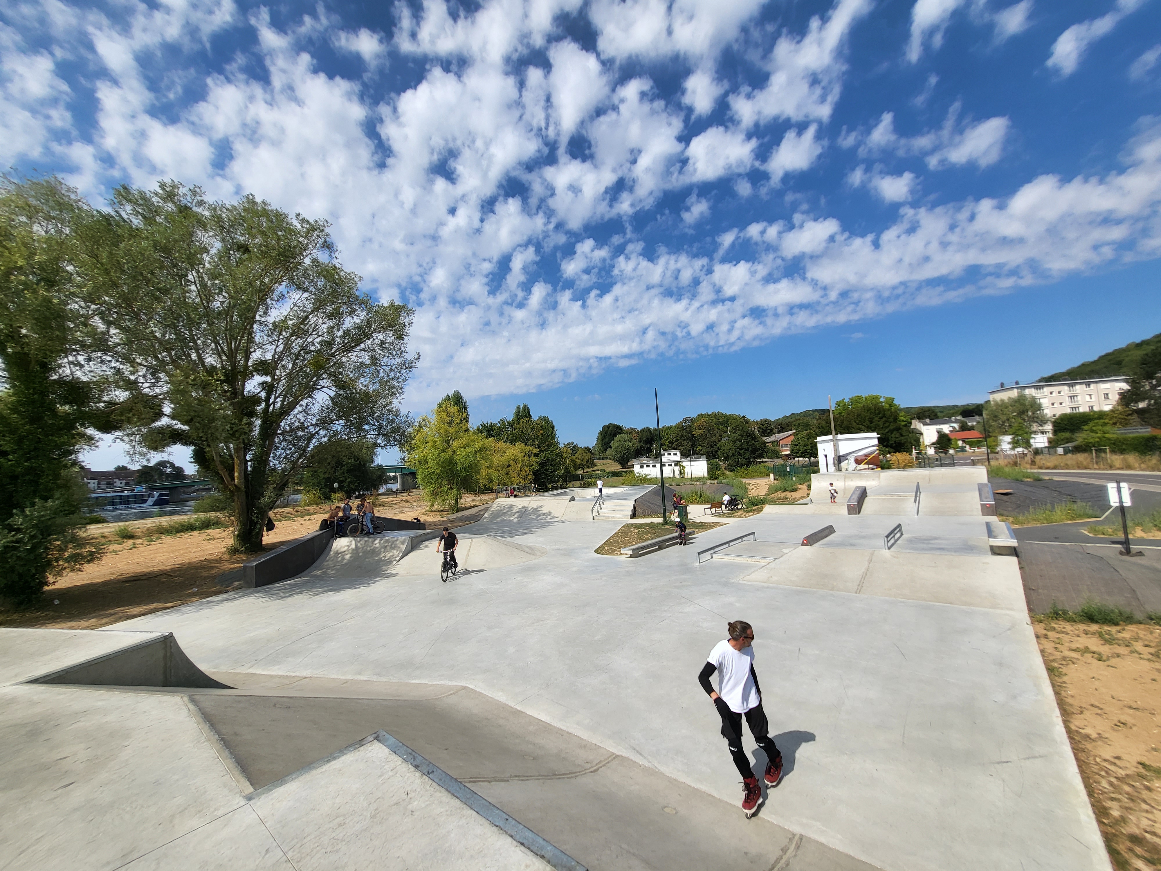 Ein weitläufiger Skatepark mit Sportlern unter einem bewölkten blauen Himmel, umgeben von Bäumen und Gebäuden.