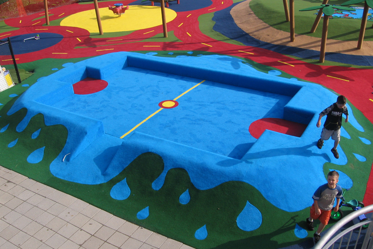 Zwei Kinder spielen auf einem farbenfrohen Spielplatz mit blauem Kletterelement in Form eines Spielfelds, umgeben von bunten Wegen und grünem Boden.