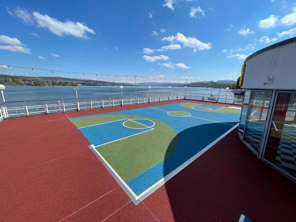 Ein Sportplatzdeck auf einem Kreuzfahrtschiff mit buntem Belag, umgeben von Wasser und blauem Himmel im Hintergrund.