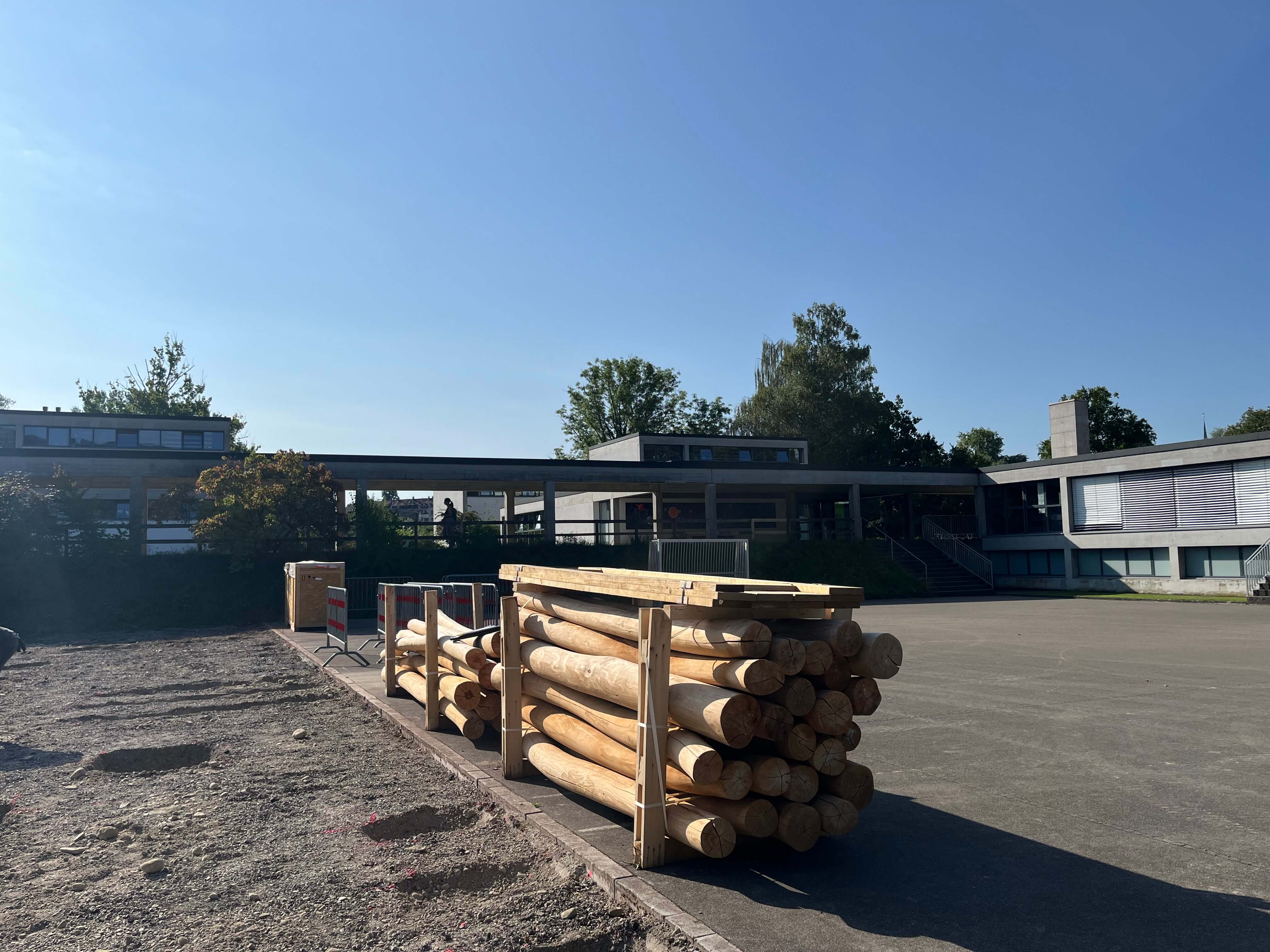 Ein Stapel Holzstämme vor einem modernen Schulgebäude mit klarem Himmel im Hintergrund