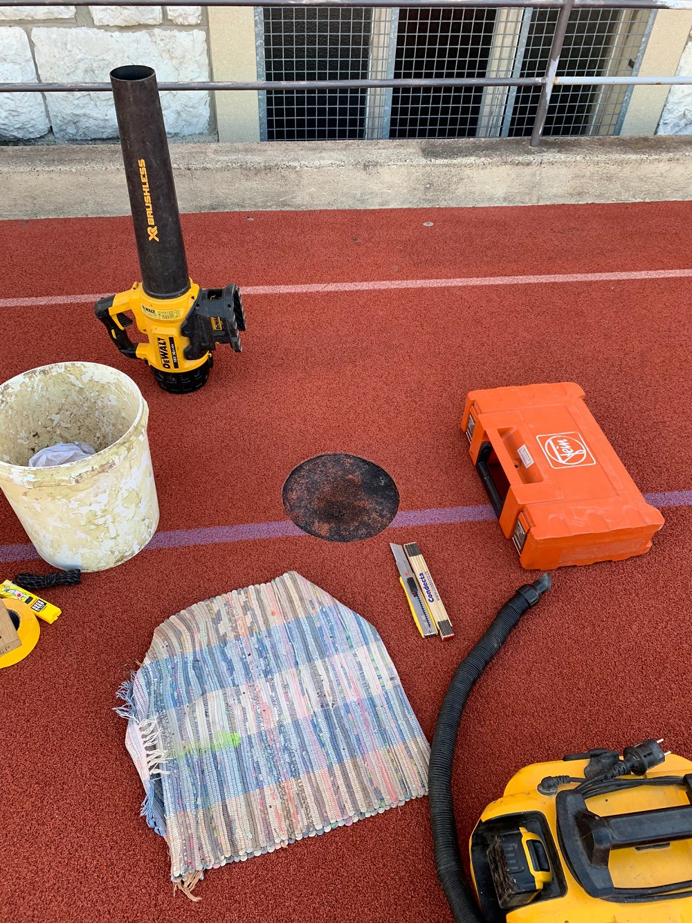 Auf einer roten Laufbahn liegen verschiedene Werkzeuge und Materialien wie ein Laubbläser, ein Staubsauger, ein Teppich, eine Wasserwaage und ein Eimer.