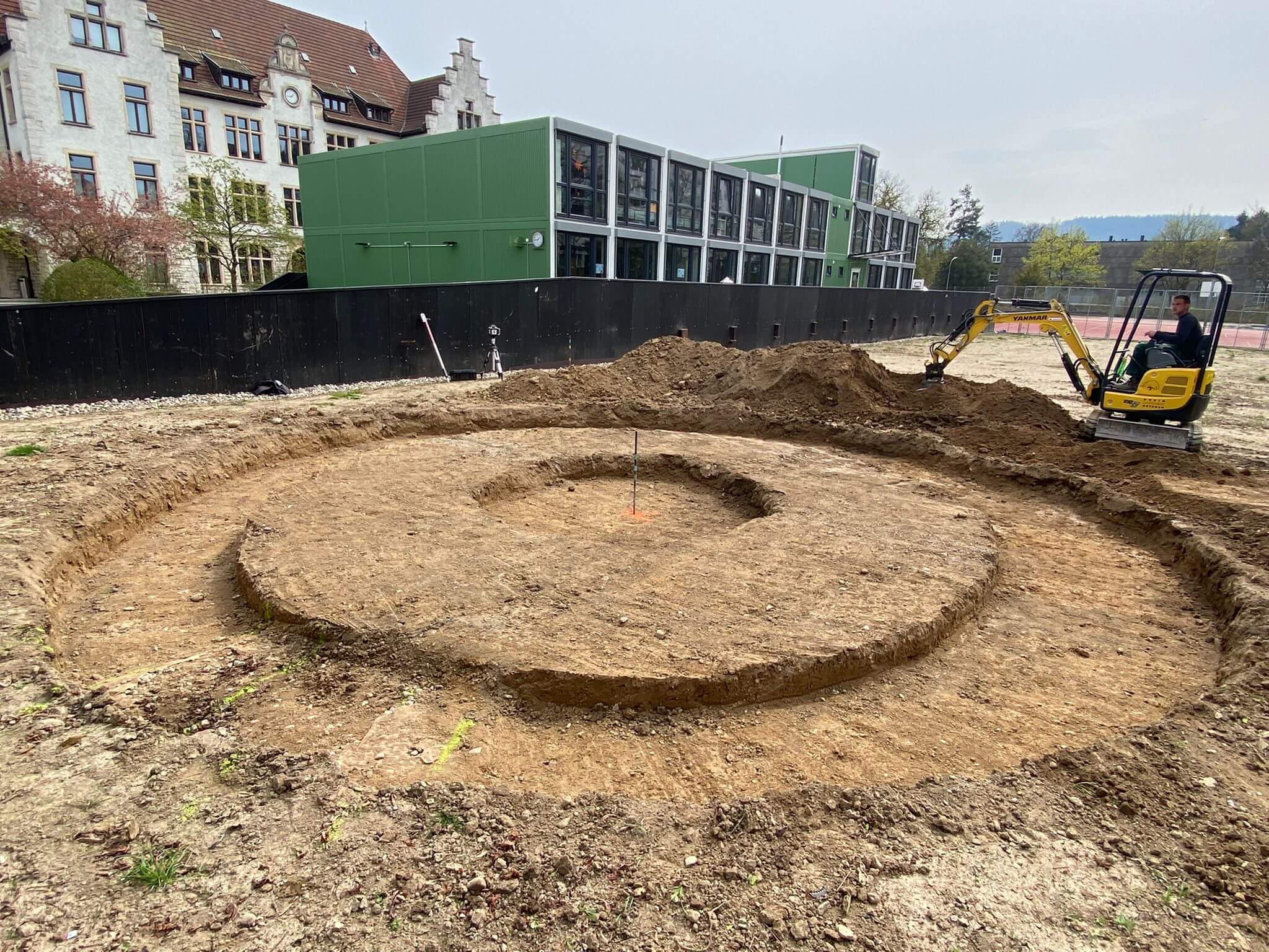 Ein Mini-Bagger bei Erdarbeiten auf einer Baustelle neben einem modernen Gebäude und einem historischen Gebäude im Hintergrund