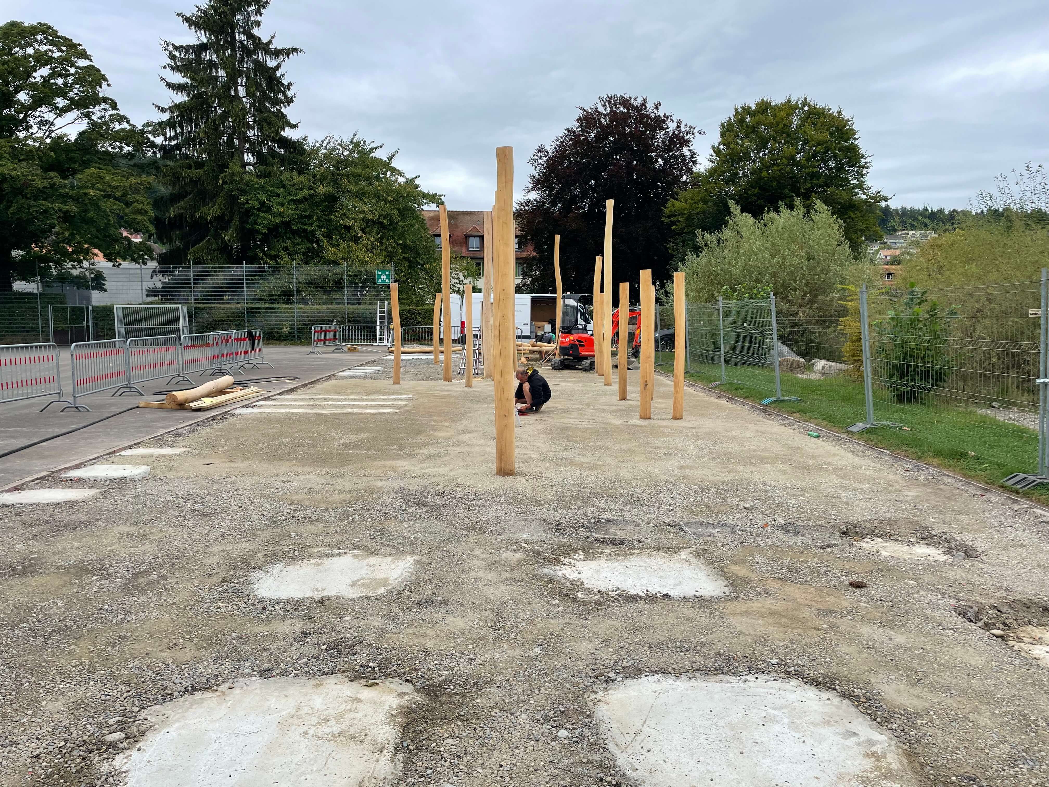 Eine Baustelle mit mehreren aufrecht stehenden Holzpfosten. Ein Bauarbeiter ist zu sehen, umgeben von Bauzäunen und Bäumen im Hintergrund.