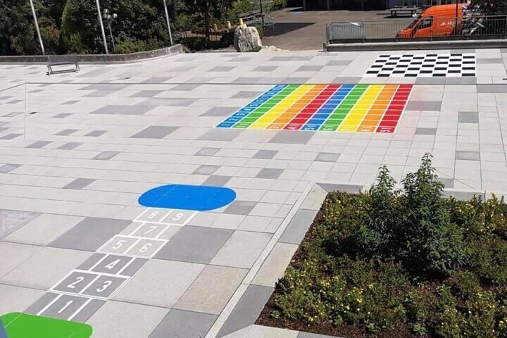 Ein gepflasterter Platz mit farbenfrohen Spielbereichen, darunter ein klassisches Hüpfspiel und ein Zahlenfeld.