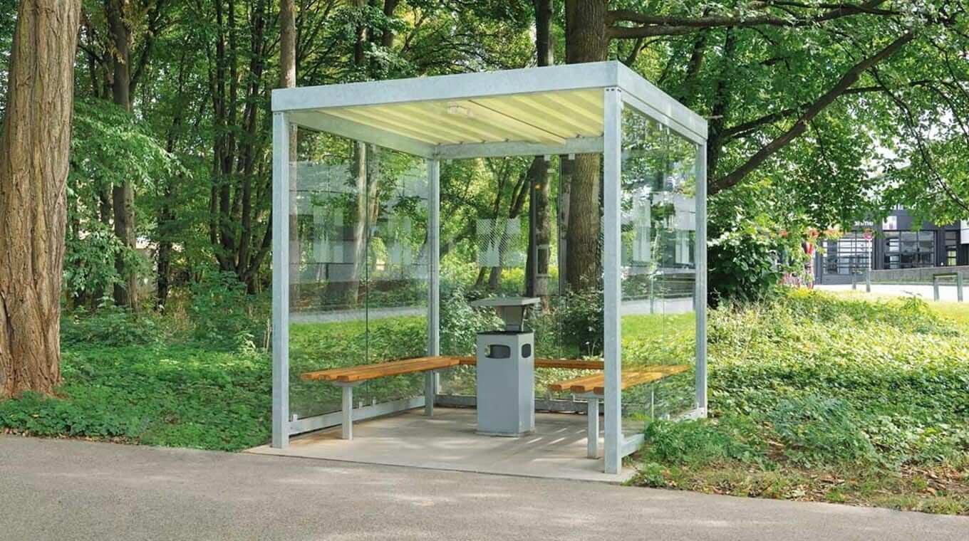 Ein moderner, überdachter Raucherunterstand aus Glas mit Sitzbänken in einem grünen Park.