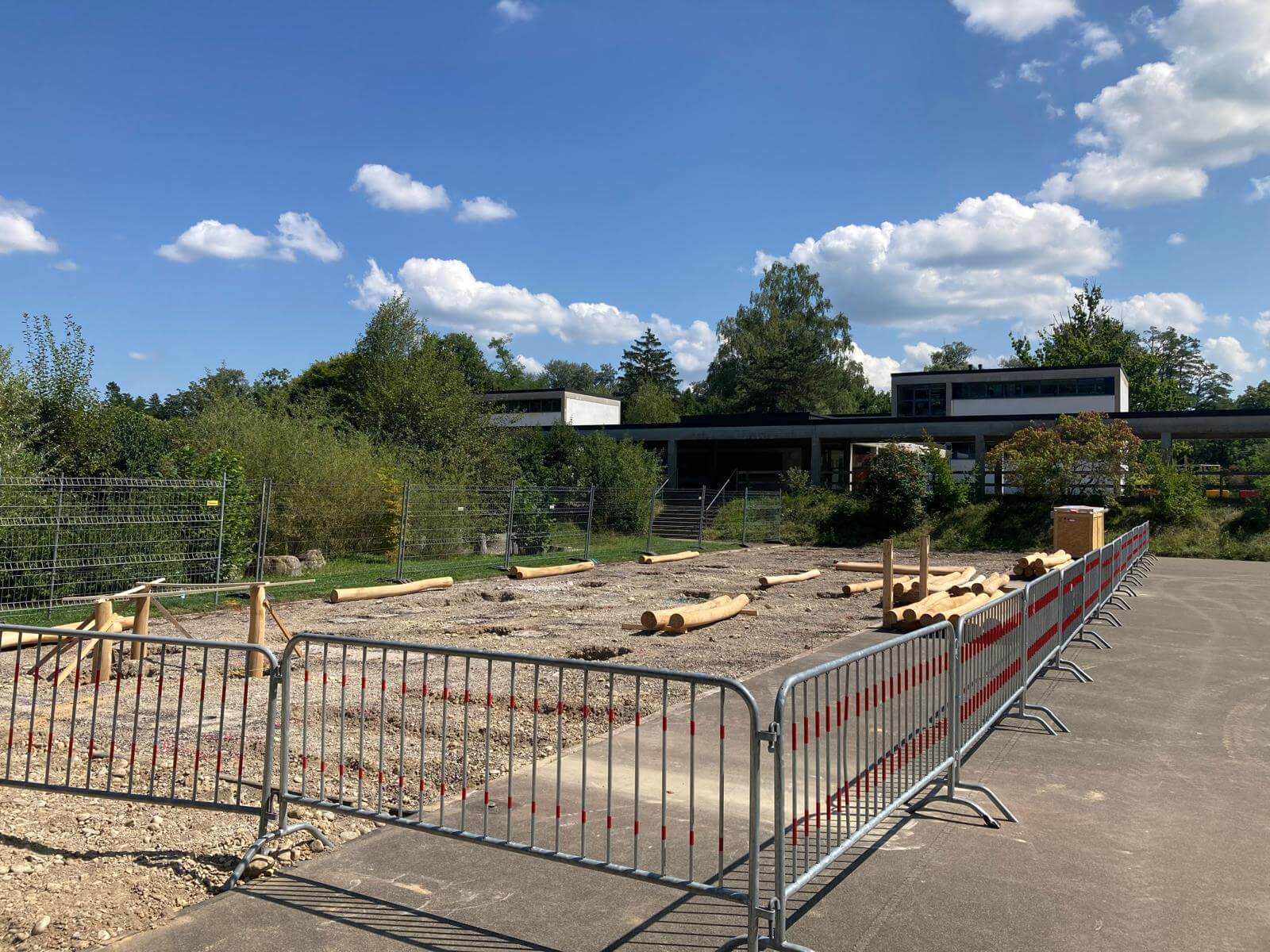 Bild einer Baustelle auf einem Schulgelände mit Absperrgittern und Holzstämmen bei blauem Himmel