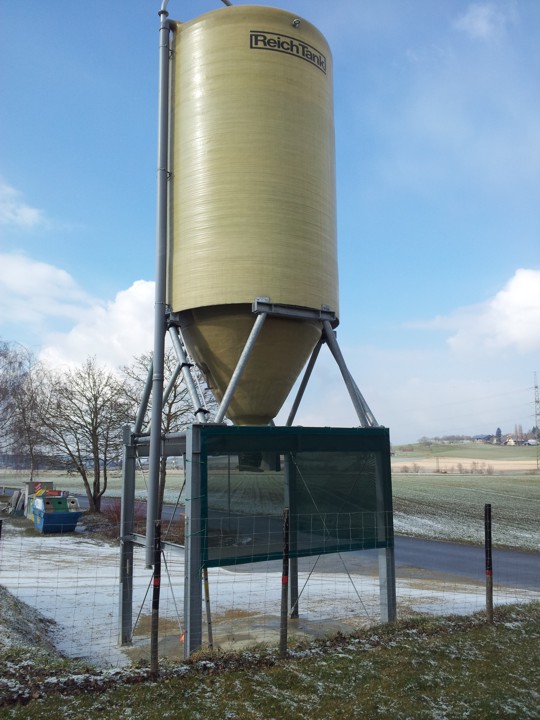 Ein großes gelbliches Silo steht auf einem verschneiten ländlichen Gebiet, umgeben von Bäumen und einer Straße.