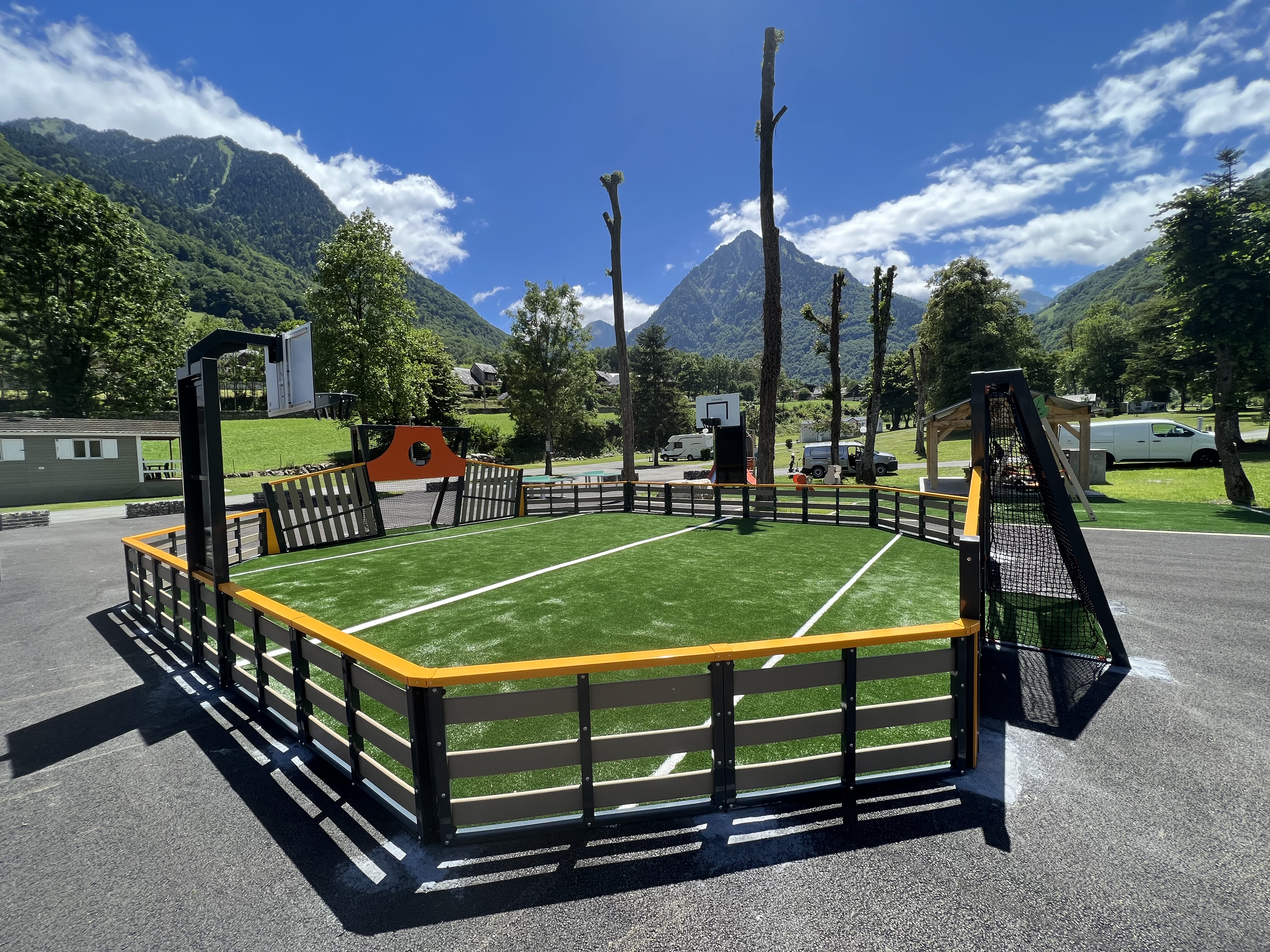 Ein moderner Multifunktions-Sportplatz mit Kunstrasen und Basketballkorb, umgeben von beeindruckender Berglandschaft und blauen Himmel.