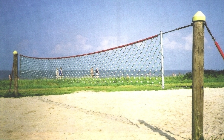Ein leeres Strandvolleyballfeld mit Netz, umgeben von Sand, grüner Wiese und Meer im Hintergrund unter einem blauen Himmel.