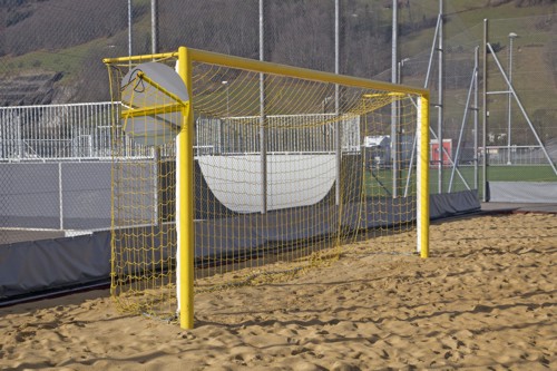 Ein gelbes Beachsoccer-Tor steht auf einem Sandplatz, umgeben von Zäunen und einem grünen Hügel im Hintergrund.