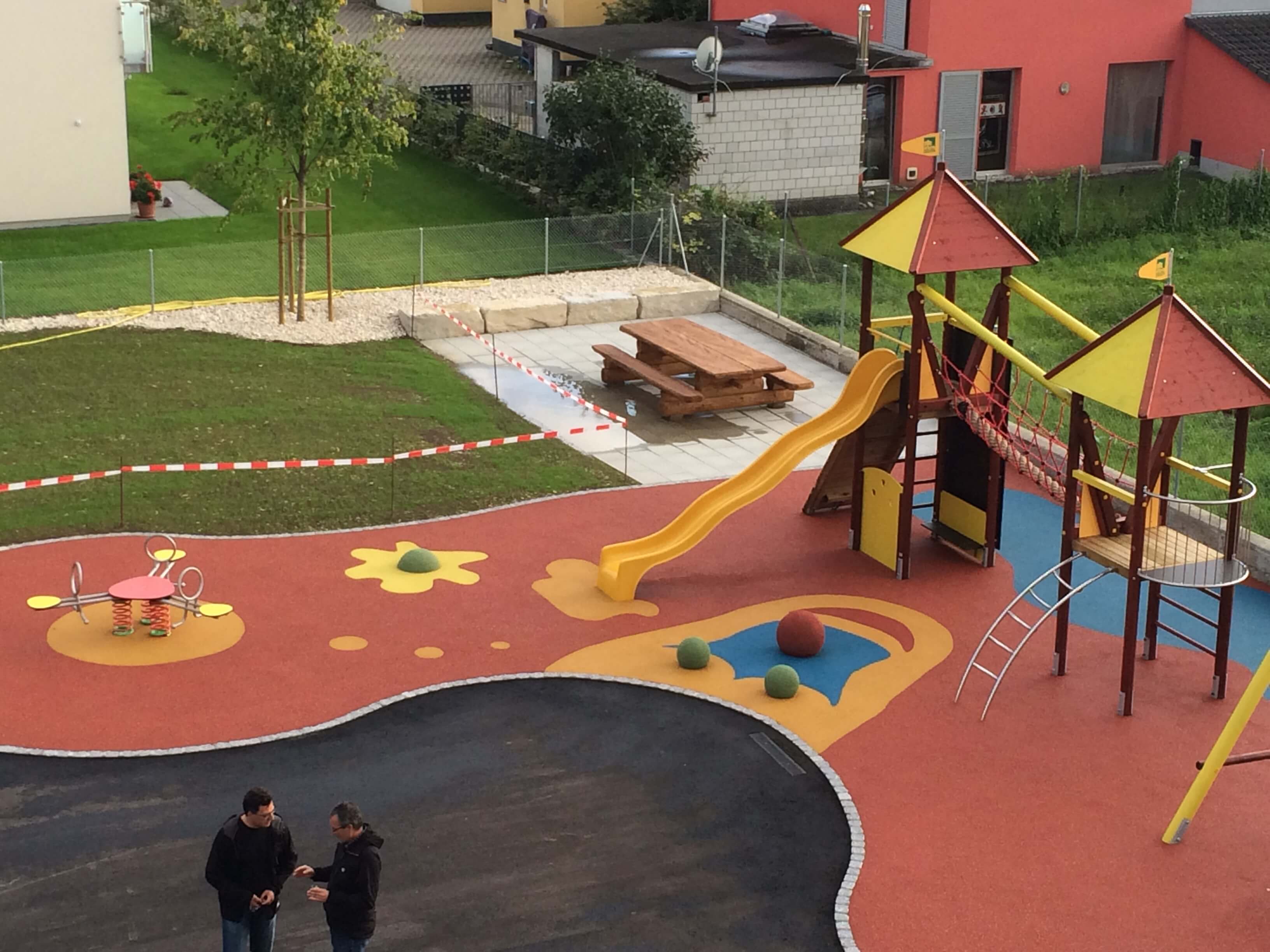 Moderner Spielplatz mit Klettergerüst, Rutsche und Wippe, umgeben von Häusern