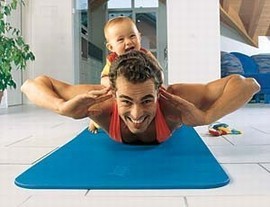Ein Vater macht Yoga auf einer blauen Matte, während sein Baby lachend auf seinem Rücken sitzt, in einem hellen Wohnzimmer