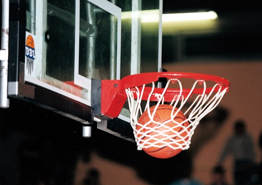Ein Basketball passiert das Netz eines Basketballkorbs in einer Sporthalle.