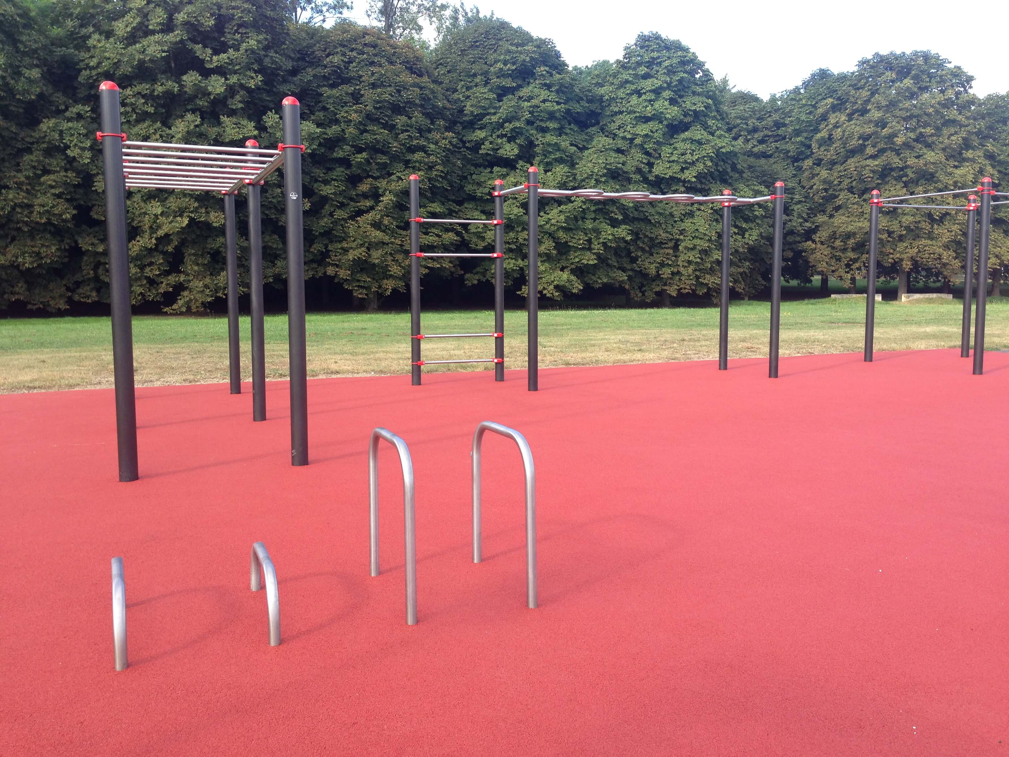 Ein moderner Outdoor-Fitnesspark mit verschiedenen Klettergeräten auf rotem Bodenbelag, umgeben von grünen Bäumen im Hintergrund.