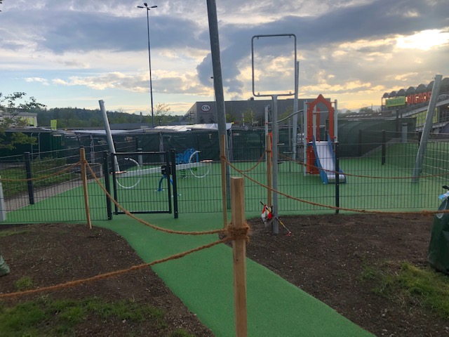 Ein moderner Kinderspielplatz im Freien mit Klettergerüst, Rutsche und grünen Bodenbelag, umgeben von einem Zaun.