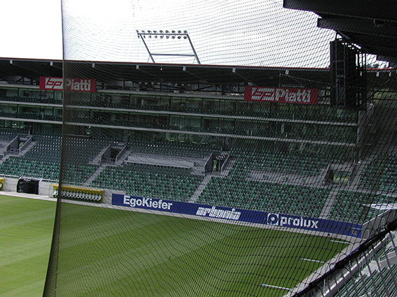 Blick auf einem leeren Fußballstadion mit grünen Tribünen, Schutznetz und Werbebannern vor einem Spiel