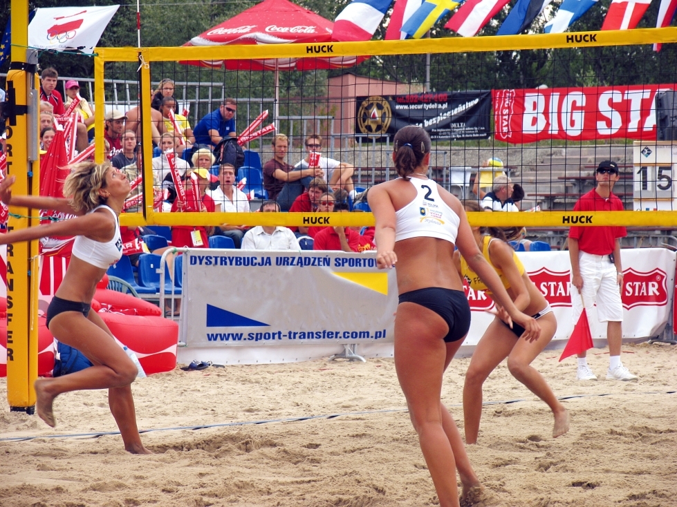 Drei Frauen beim Beachvolleyballspiel auf einem Sandplatz während eines Turniers, umgeben von Zuschauern und Bannern
