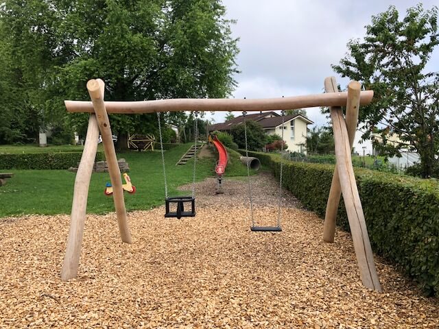Ein einladender Spielplatz mit Holzschaukel, Rutsche und Hackschnitzeluntergrund, umgeben von Bäumen und Rasen.