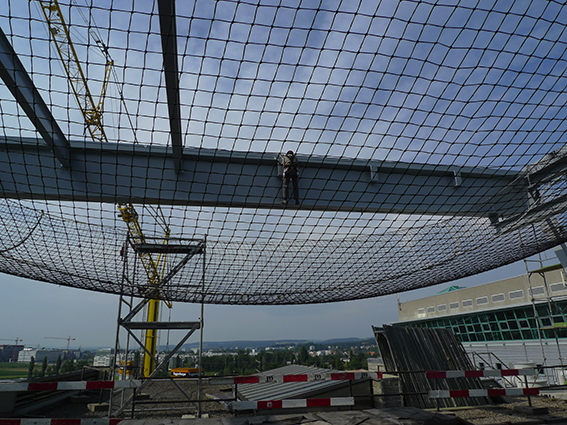 Ein Bauarbeiter steht auf einem Stahlträger über einem Sicherheitsnetz an einer Baustelle mit Kran im Hintergrund.