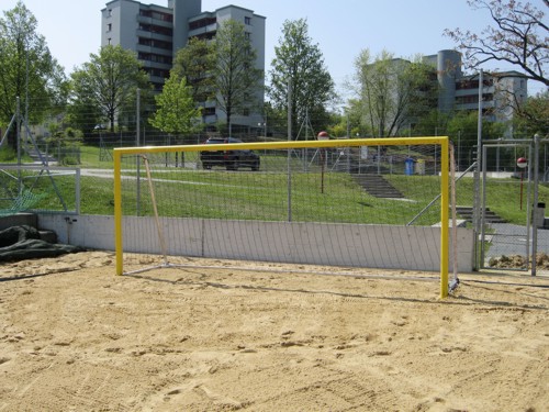 Ein gelbes Fußballtor auf einem Sandplatz, umgeben von Bäumen und Wohnhäusern
