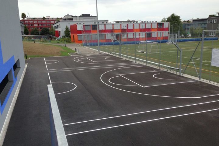 Ein moderner Schulhof mit einem neu angelegten Basketballplatz und einem angrenzenden Fußballfeld. Im Hintergrund sind Schulgebäude zu sehen.
