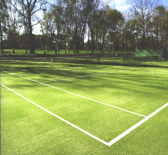 Ein leerer grüner Tennisplatz im Freien, umgeben von Bäumen, mit Netz im Vordergrund und klarer Himmel im Hintergrund.