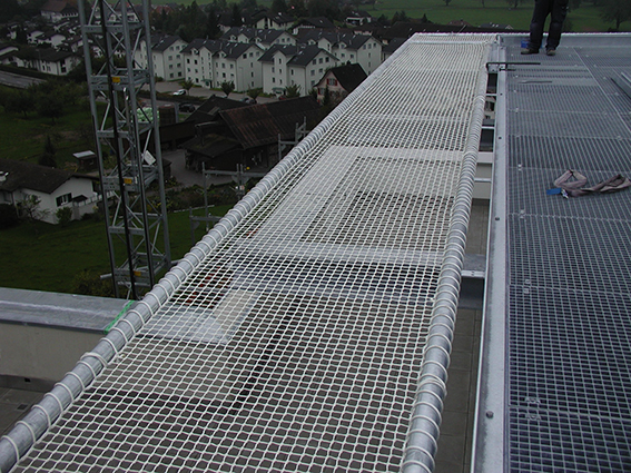 Ein Sicherheitsnetz an einer Gebäudefassade während Bauarbeiten, mit einem Ausblick auf eine ländliche Umgebung im Hintergrund.