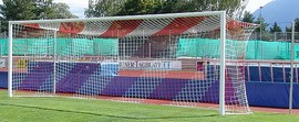 Un but de football sur un terrain vert avec une tribune colorée en arrière-plan, entouré d'arbres et d'un ciel ensoleillé.