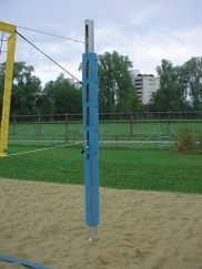 Ein verstellbarer Volleyballpfosten auf einem Sandplatz mit grünem Hintergrund und bewölktem Himmel.