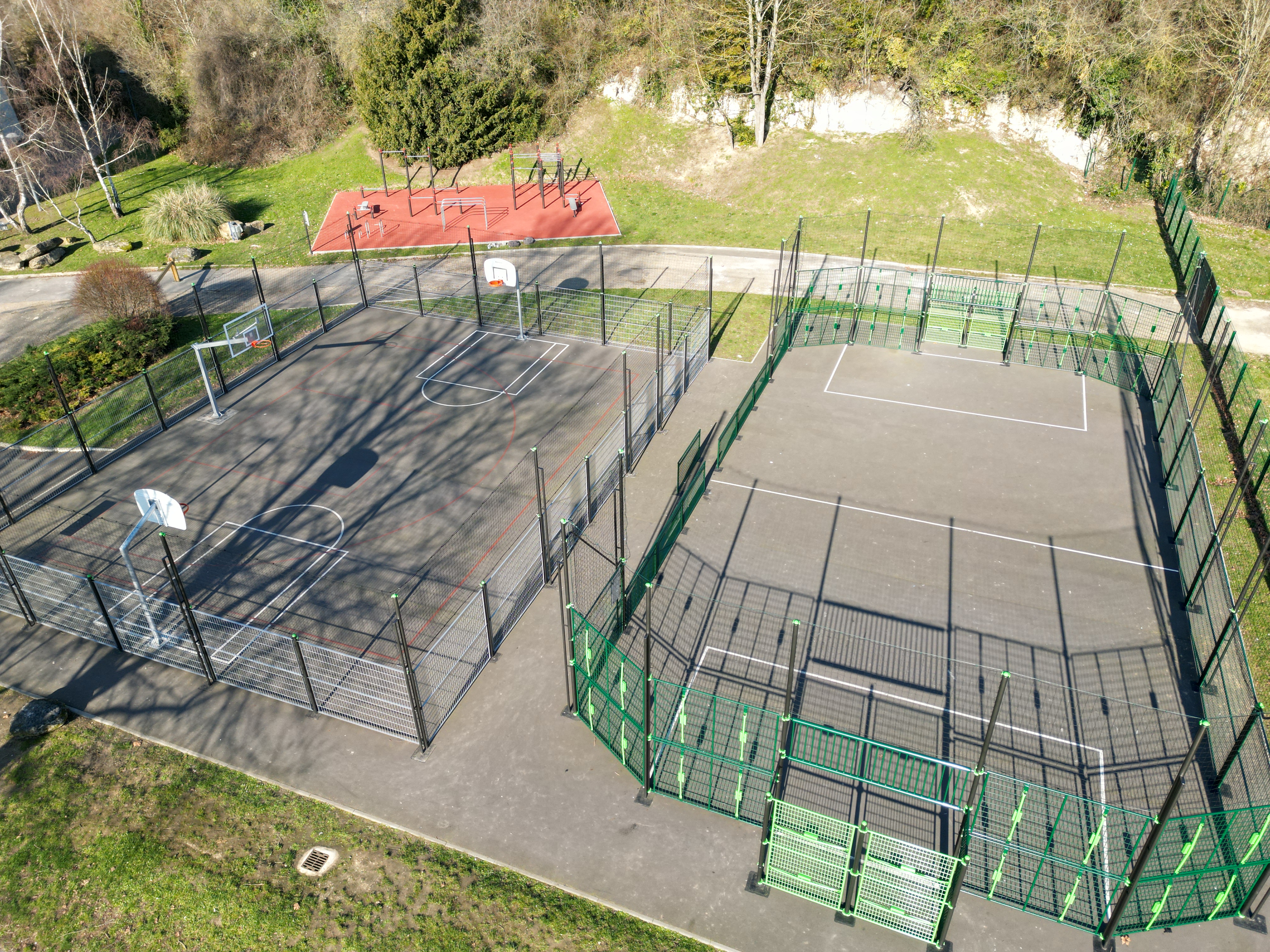 Luftaufnahme eines Parks mit zwei eingezäunten Sportplätzen, einem Basketballplatz und einem Mehrzweckplatz, umgeben von Bäumen und Grünflächen.